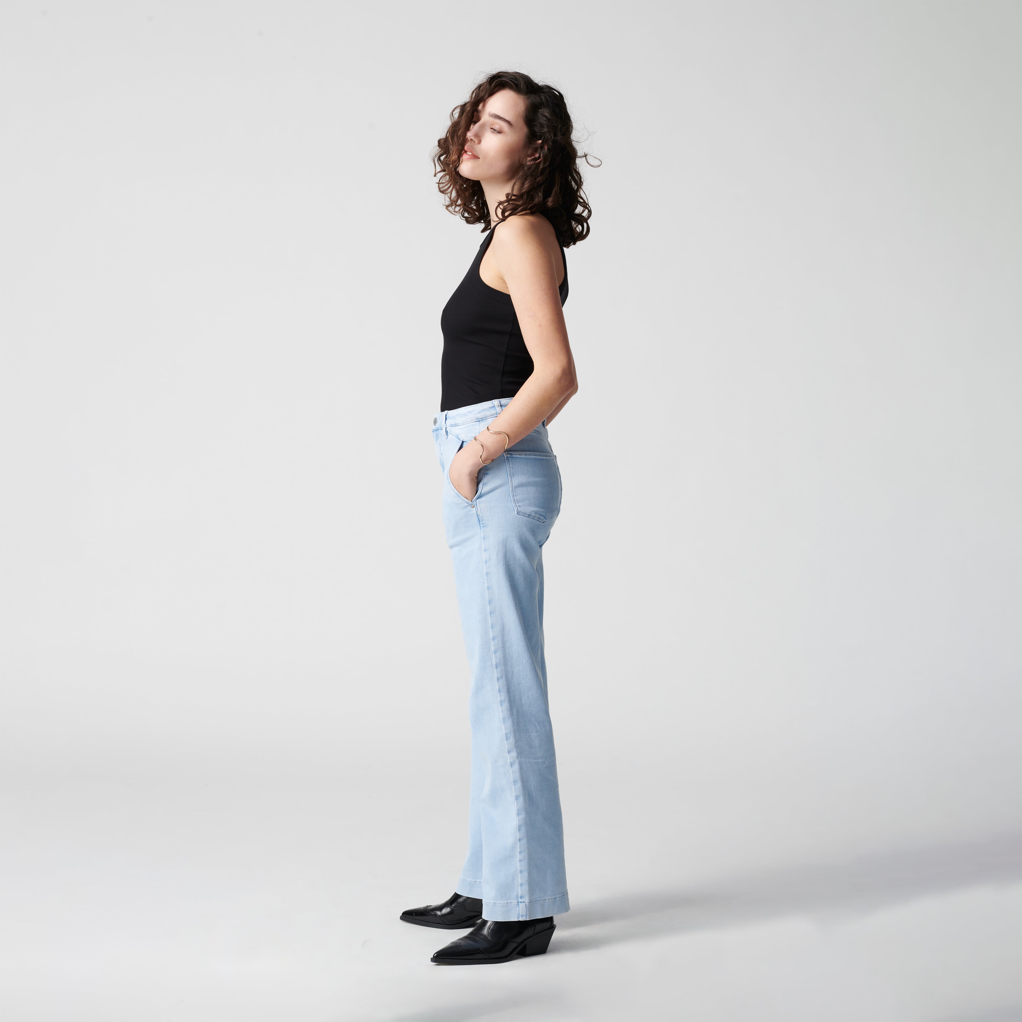 Seitenansicht einer Frau in hellblaue High-Waist-Flare-Jeans mit französischen Taschen (DEW Flared Soft Denim, Bright Blue) und schwarzem Top.