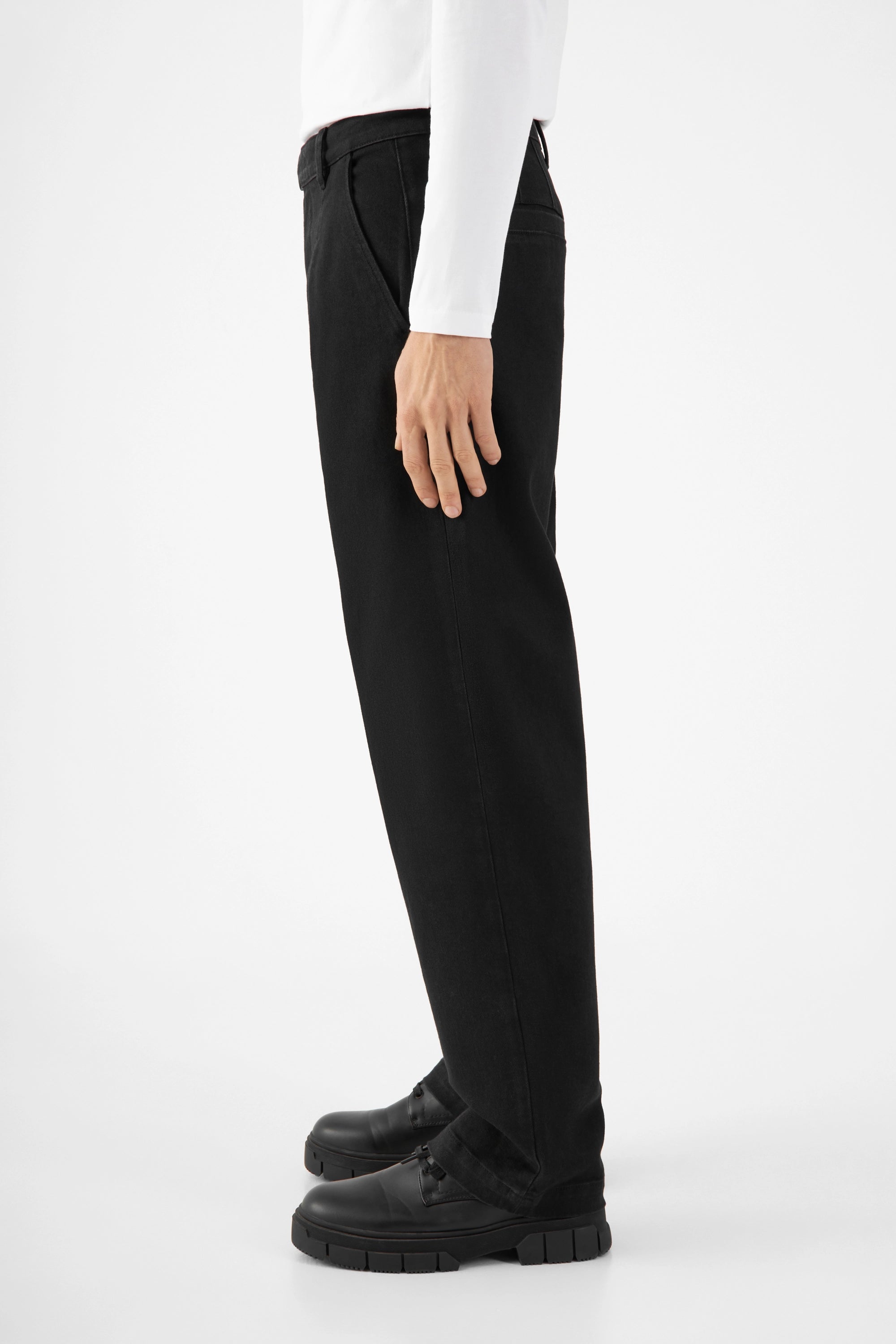 Seitliche Ansicht einer schwarzen ASTER Balloon-Soft-TENCEL Hose im Balloon-Fit: mittlerer Bund, weite Hüfte/Oberschenkel, zum Saum hin schmal, getragen mit weißem Hemd und schwarzen Schuhen.