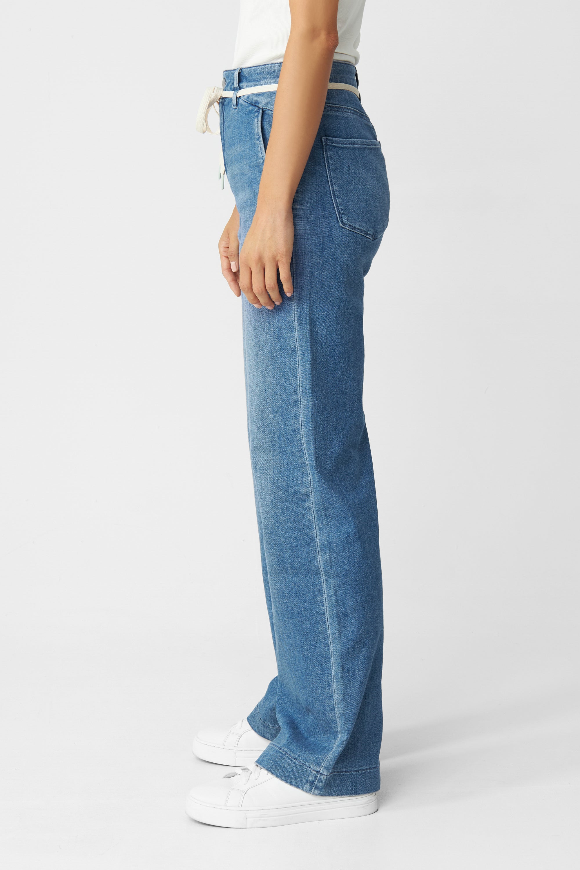 Seitenansicht der DEW Flared Soft Denim in Medium Blue: hohe Taille, ausgestelltes Bein, französische Taschen; getragen mit weißen Sneakern.