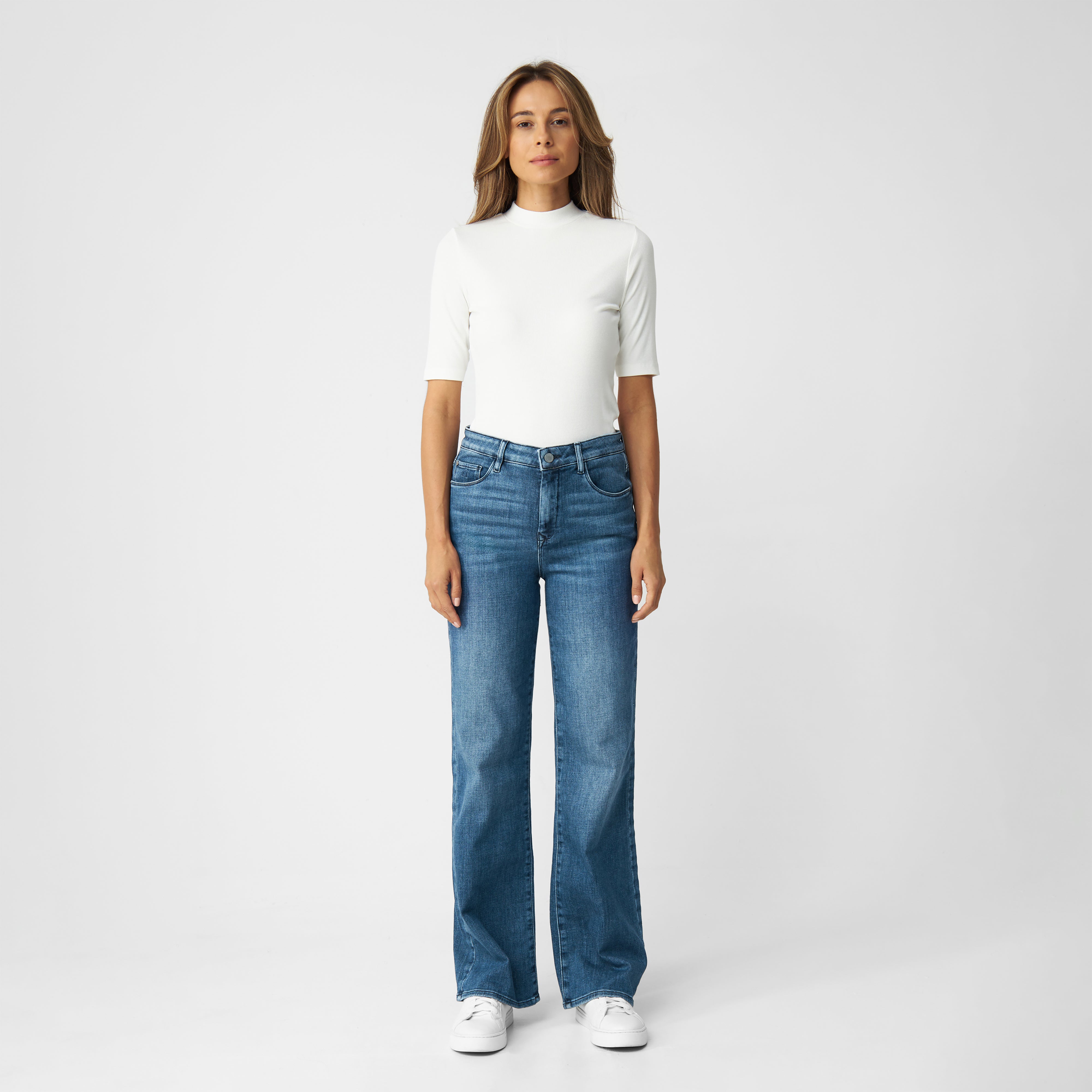 DEW Flared Soft Denim – Classic, Medium Blue: High-Waist, ausgestelltes Bein, Five-Pocket-Design, Model in weißem Oberteil.