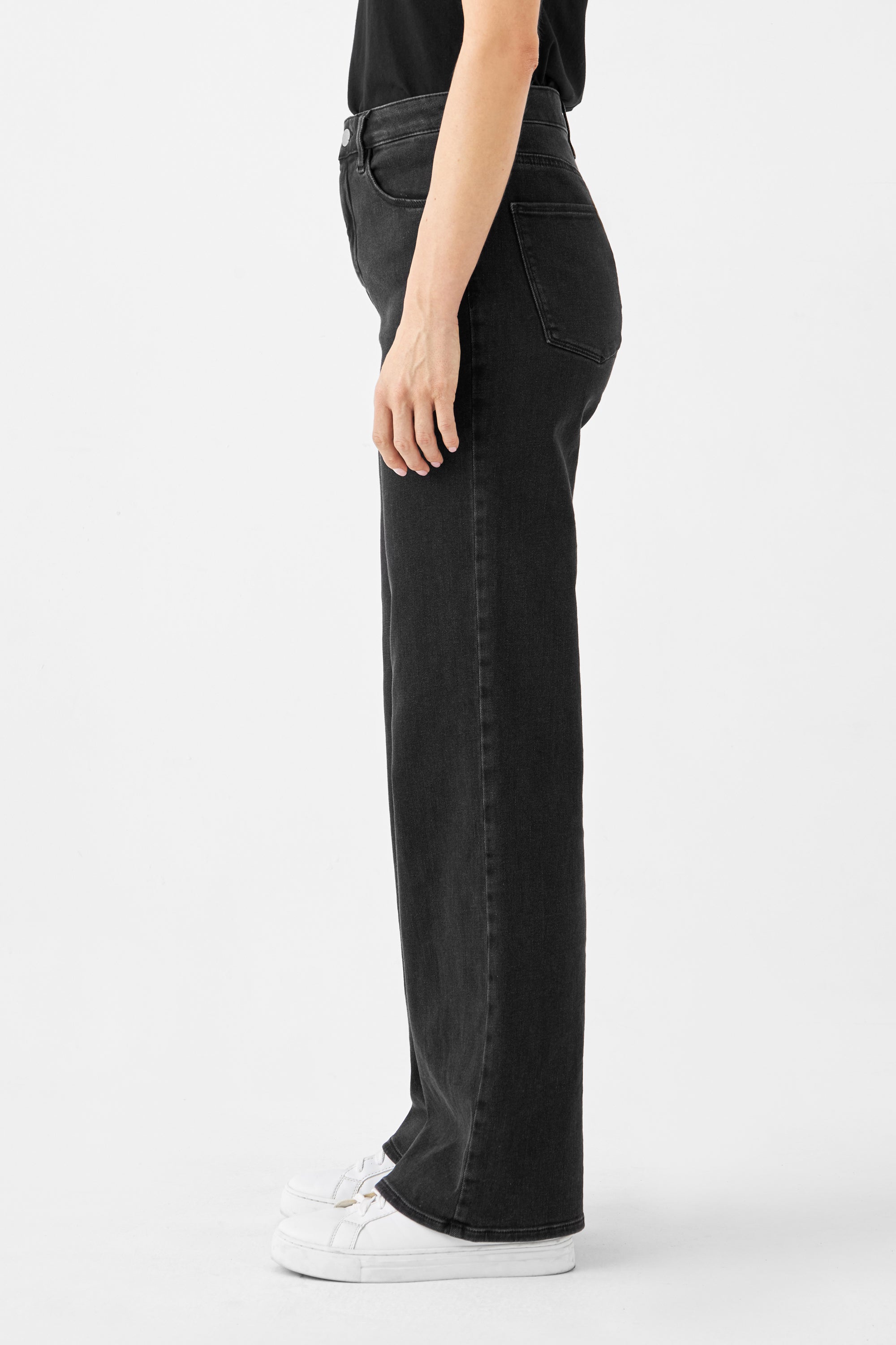 Seitliche Ansicht einer schwarzen DEW Classic High-Waist Jeans mit ausgestelltem Bein und Five-Pocket-Design, kombiniert mit weißen Sneakern.