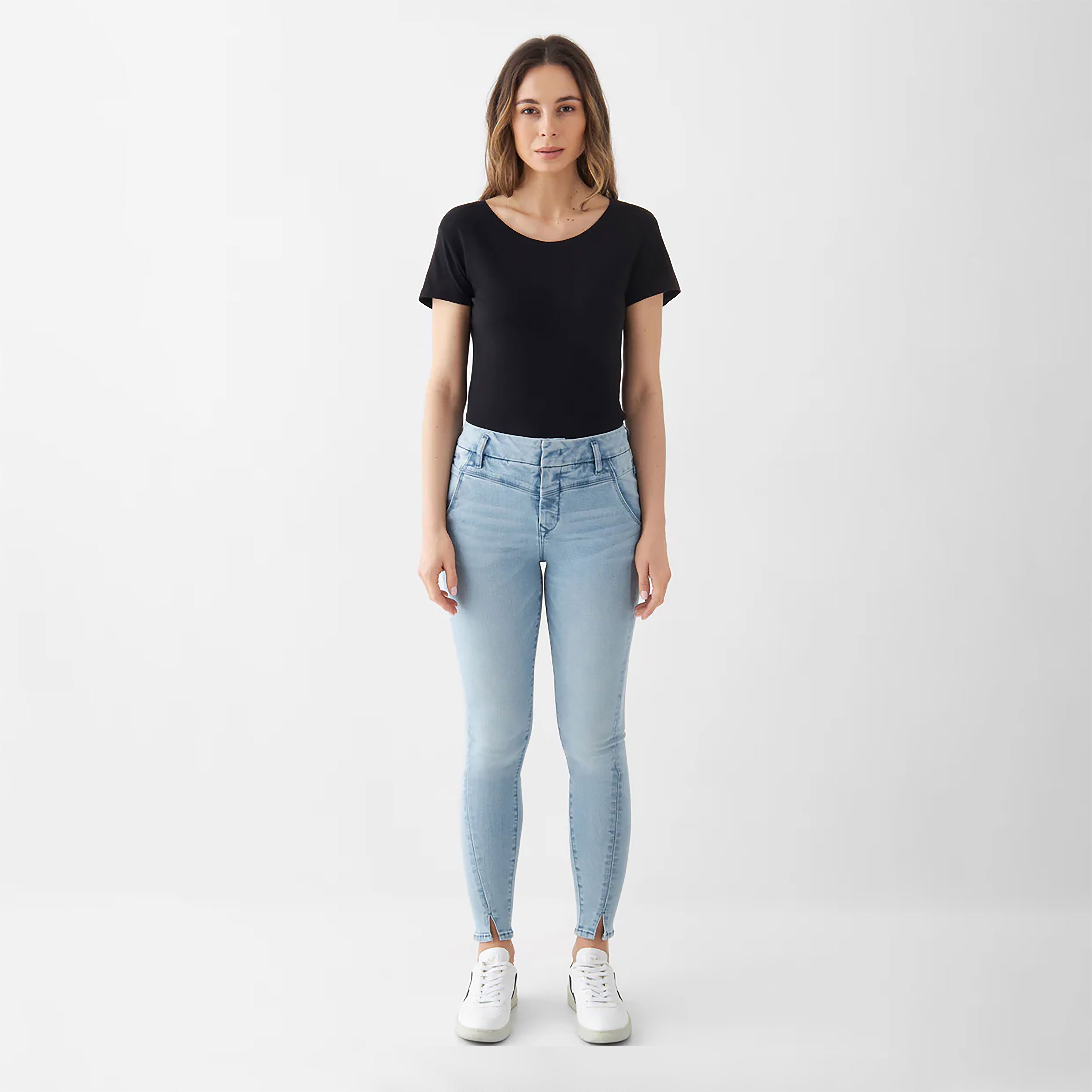 Sun Up Skinny Jeans in Light Blue; schräger Außennaht und Saumschlitz, getragen von einer Frau in schwarzem Top und weißen Sneakern.