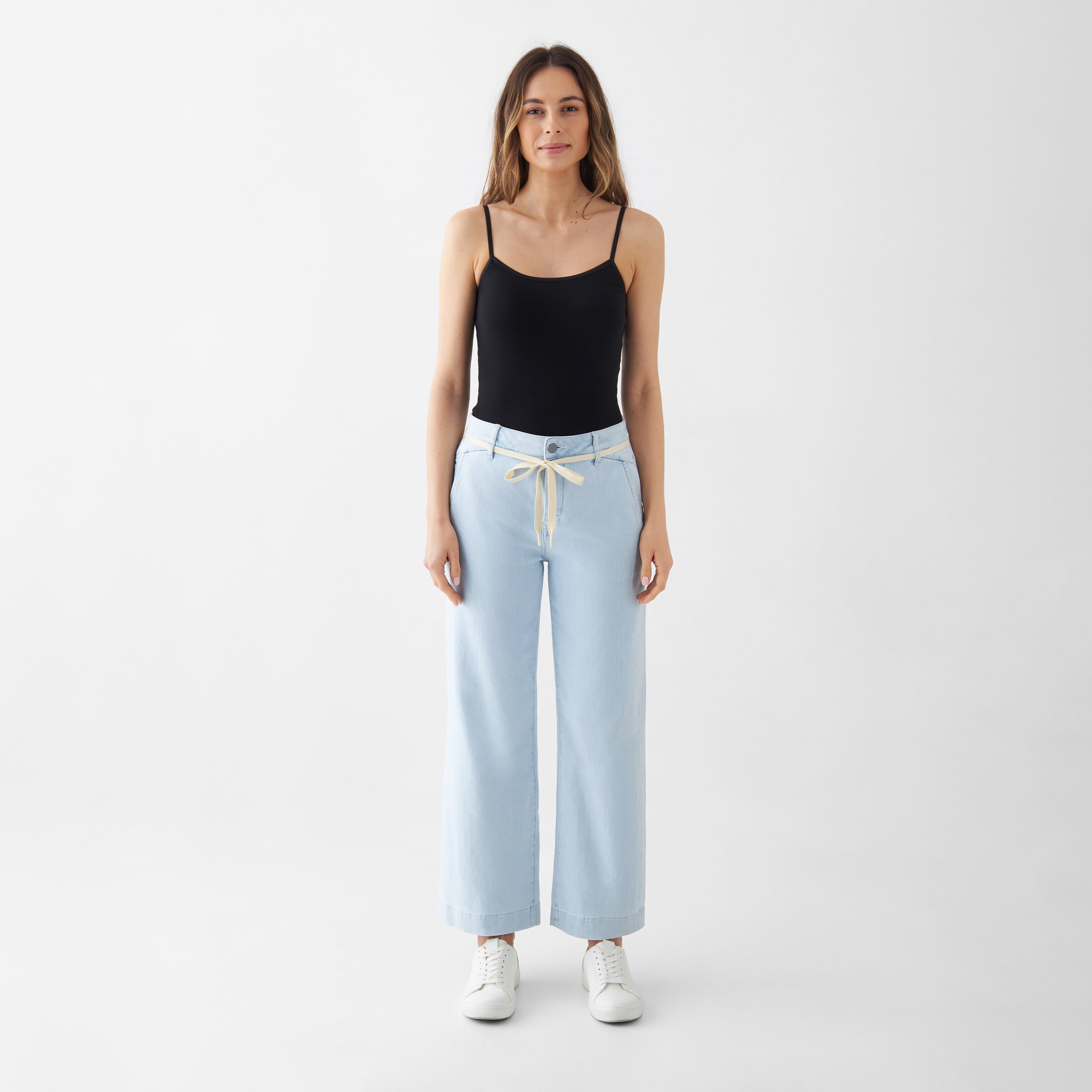hellblaues, hoch tailliertes DEW Cropped Jeans mit ausgestelltem Bein und französischen Taschen; beiger Kordelgürtel, kombiniert mit schwarzem Top und weißen Sneakern.