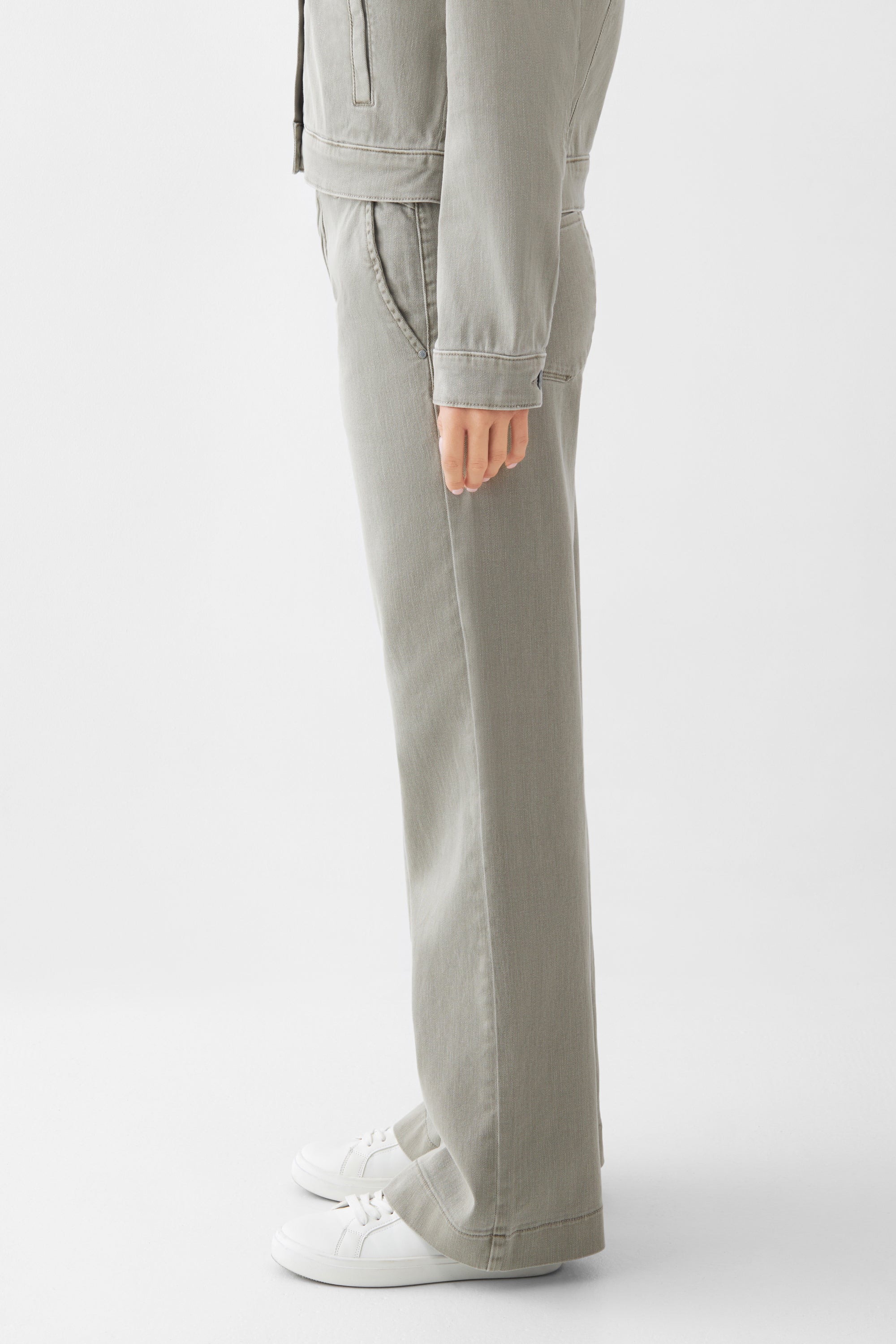 Seitliche Ansicht einer Rock Olive DEW French Pocket High-Waist Jeans mit ausgestelltem Bein aus Soft Denim; französische Taschen, weiße Sneaker.