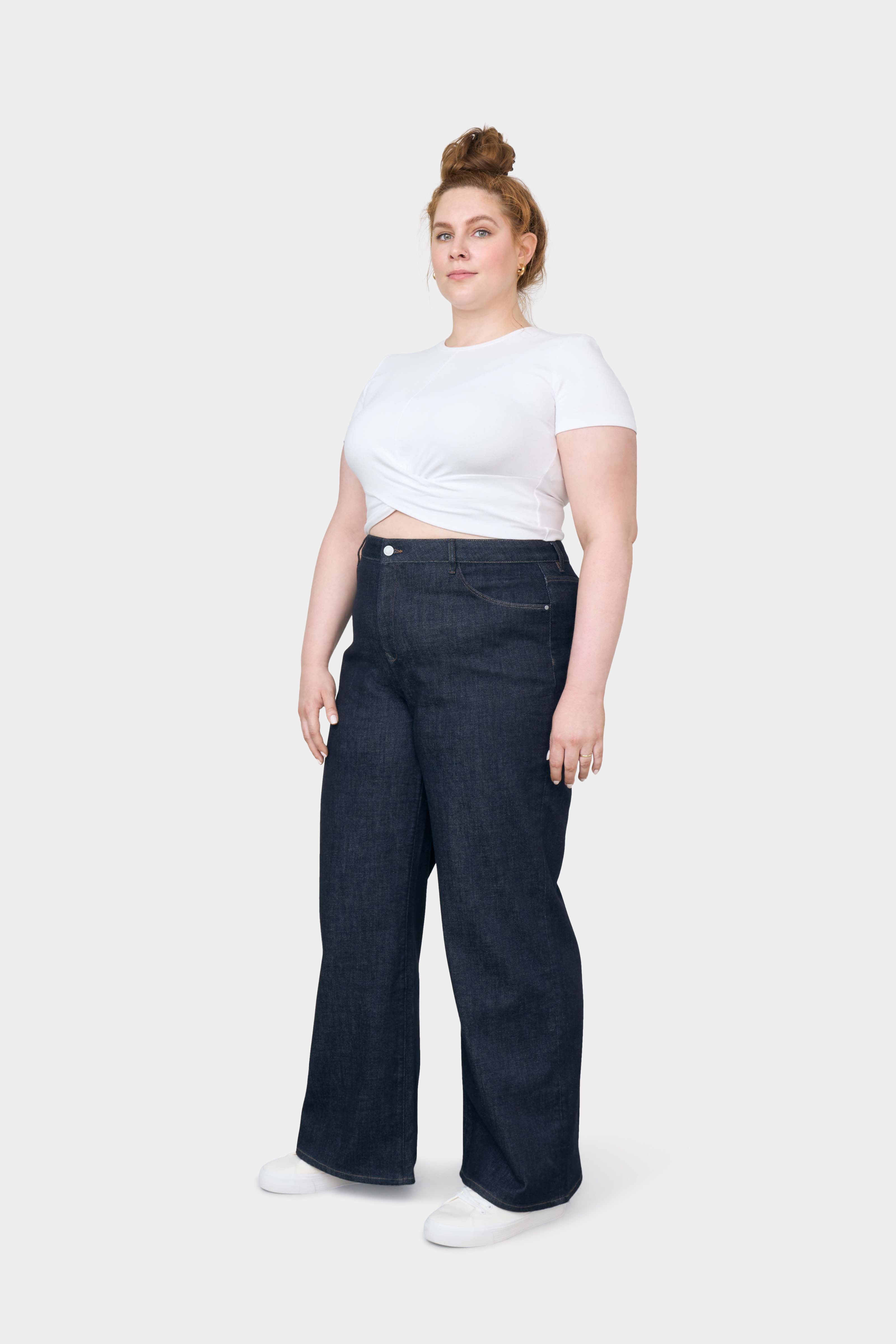 DEW Flared Soft Denim in Raw Blue: High-Waist, ausgestelltes Bein, klassisches Five-Pocket-Design; Model trägt ein weißes, vorne geknotetes T‑Shirt und weiße Sneaker.