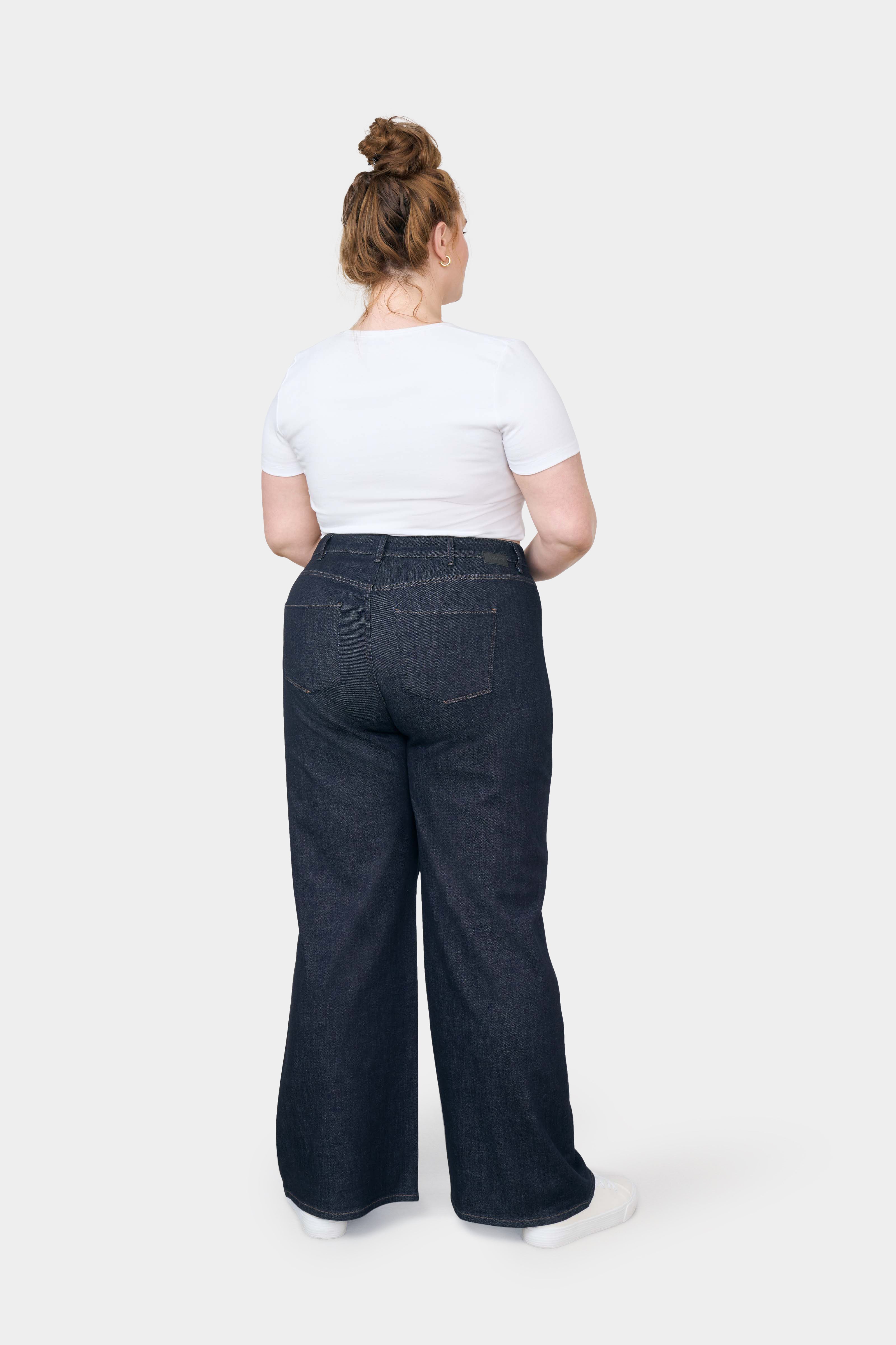 Rückansicht einer curvy Frau in dunkler Raw Blue High-Waist Jeans mit ausgestelltem Flare-Bein, weißes T‑Shirt, Sneaker.