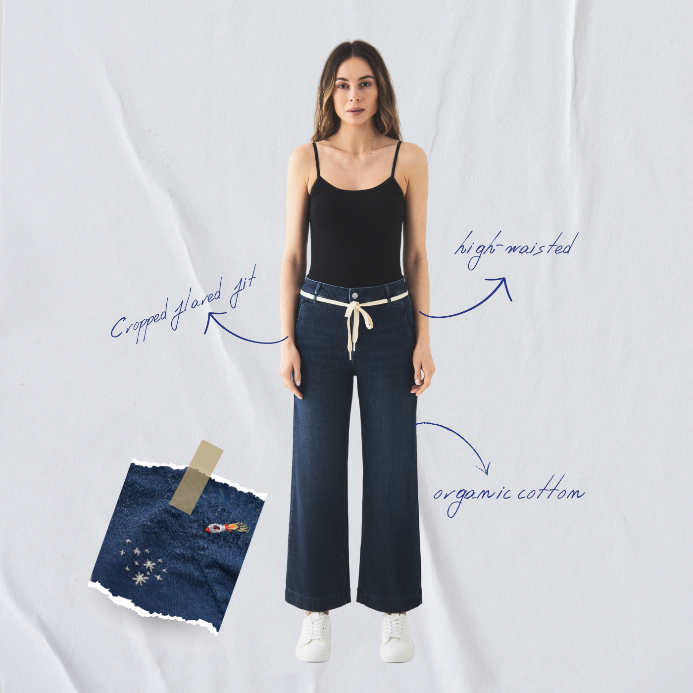 Modell trägt dunkelblaue DEW Cropped French Pocket Jeans, hoch tailliert, ausgestelltes Bein, mit hellbeigem Gürtel; schwarzes Top, weiße Sneaker; Patch‑Detail Broken Pieces sichtbar.