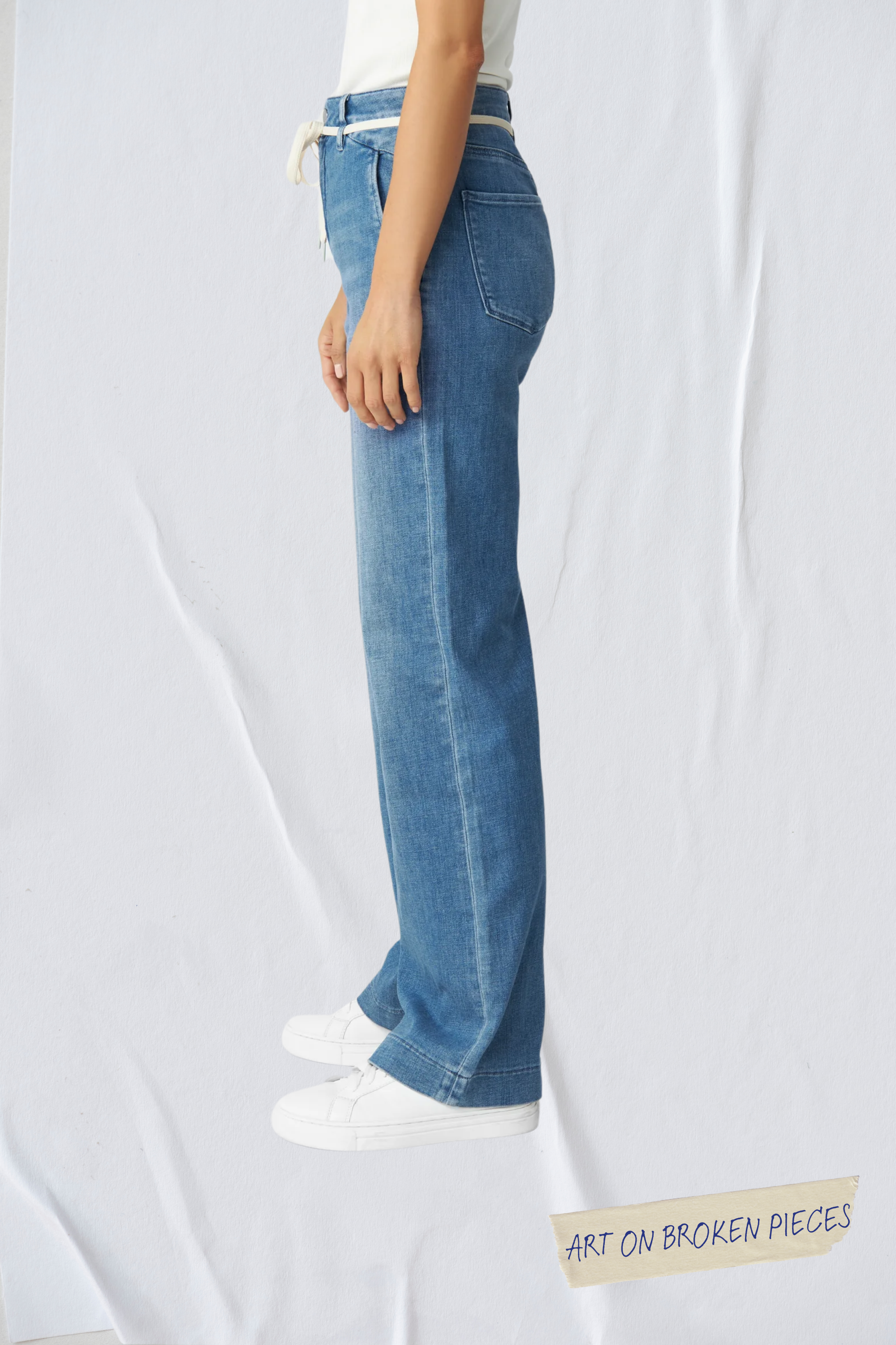 Seitliche Ansicht der DEW Flared Soft Denim Jeans in Medium Blue – hohe Taille, ausgestelltes Bein, französische Taschen; weißer Gürtel, Art on Broken Pieces.