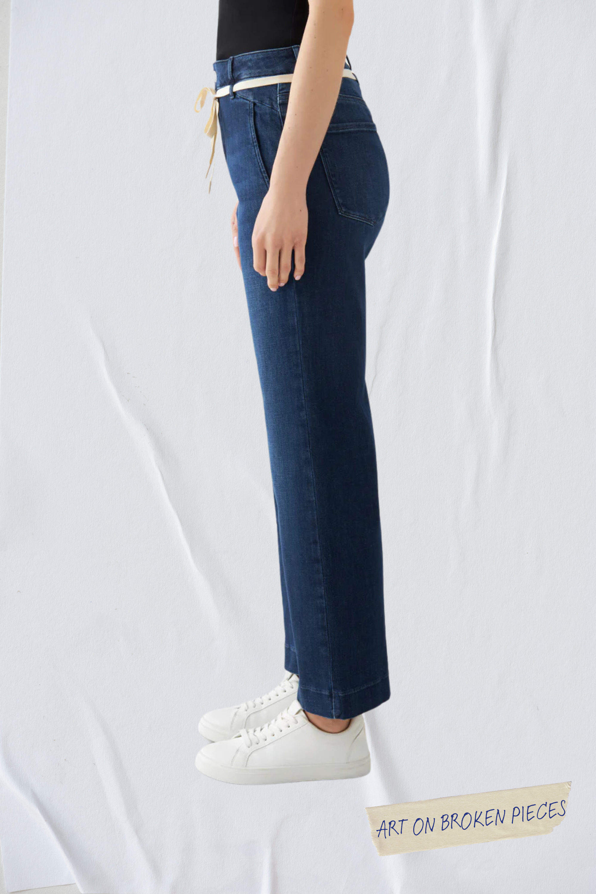 Seitenansicht einer dunkelblauen, hoch taillierten Cropped Flared Jeans mit ausgestelltem Bein, hellem Gürtel und weißen Sneakern.