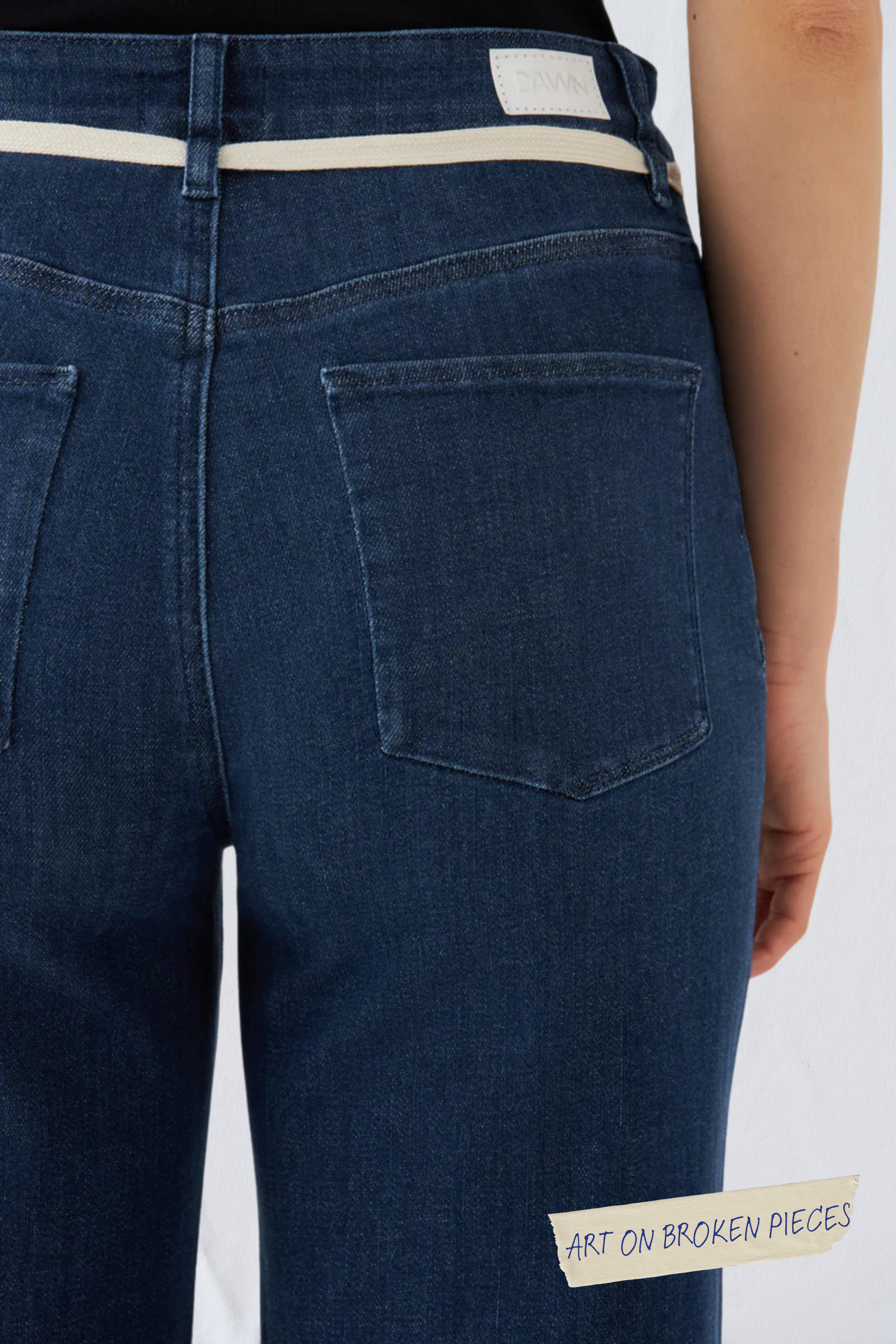 Rückansicht einer dunkelblauen DEW Flared Soft Denim Jeans mit französischen Taschen, weißem Bund-Label und dem 'Art on Broken Pieces'-Detail.