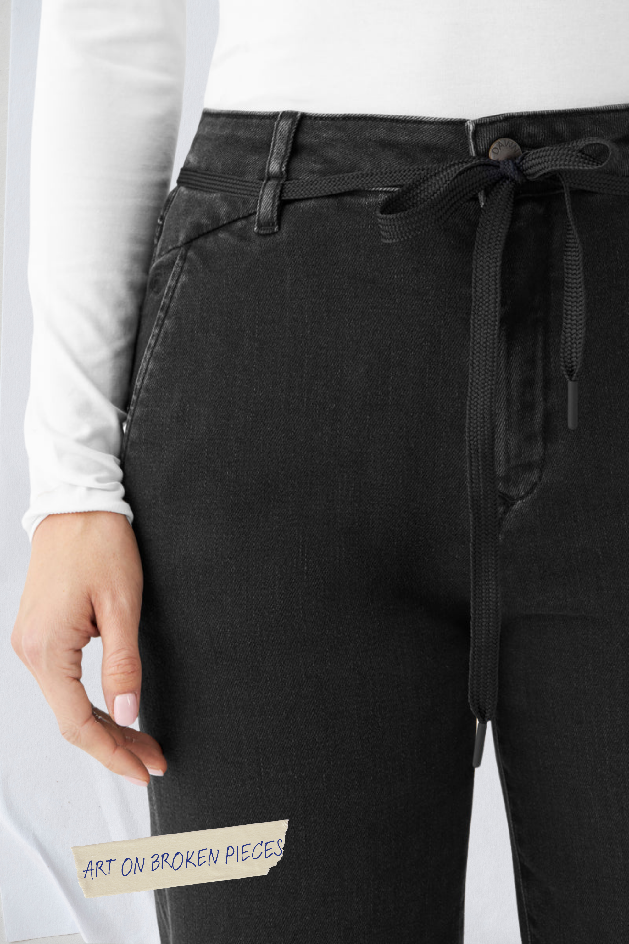 Nahaufnahme einer schwarzen DEW Jeans im Flared-Schnitt mit hohem Taillenbund und französischen Taschen; Bund mit Kordelzug und Aufkleber ART ON BROKEN PIECES am Bein.