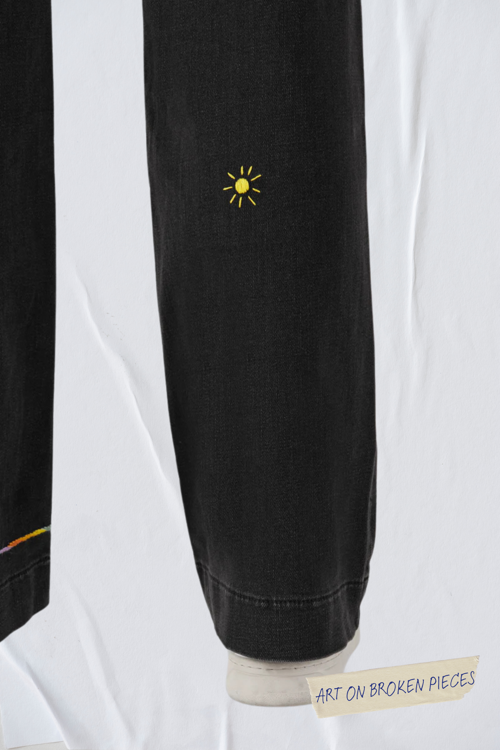 Schwarze DEW Flared Jeans im French Pocket-Stil; Bein mit gelber Sonnenstickerei und Label 'Art on Broken Pieces' am Saum.