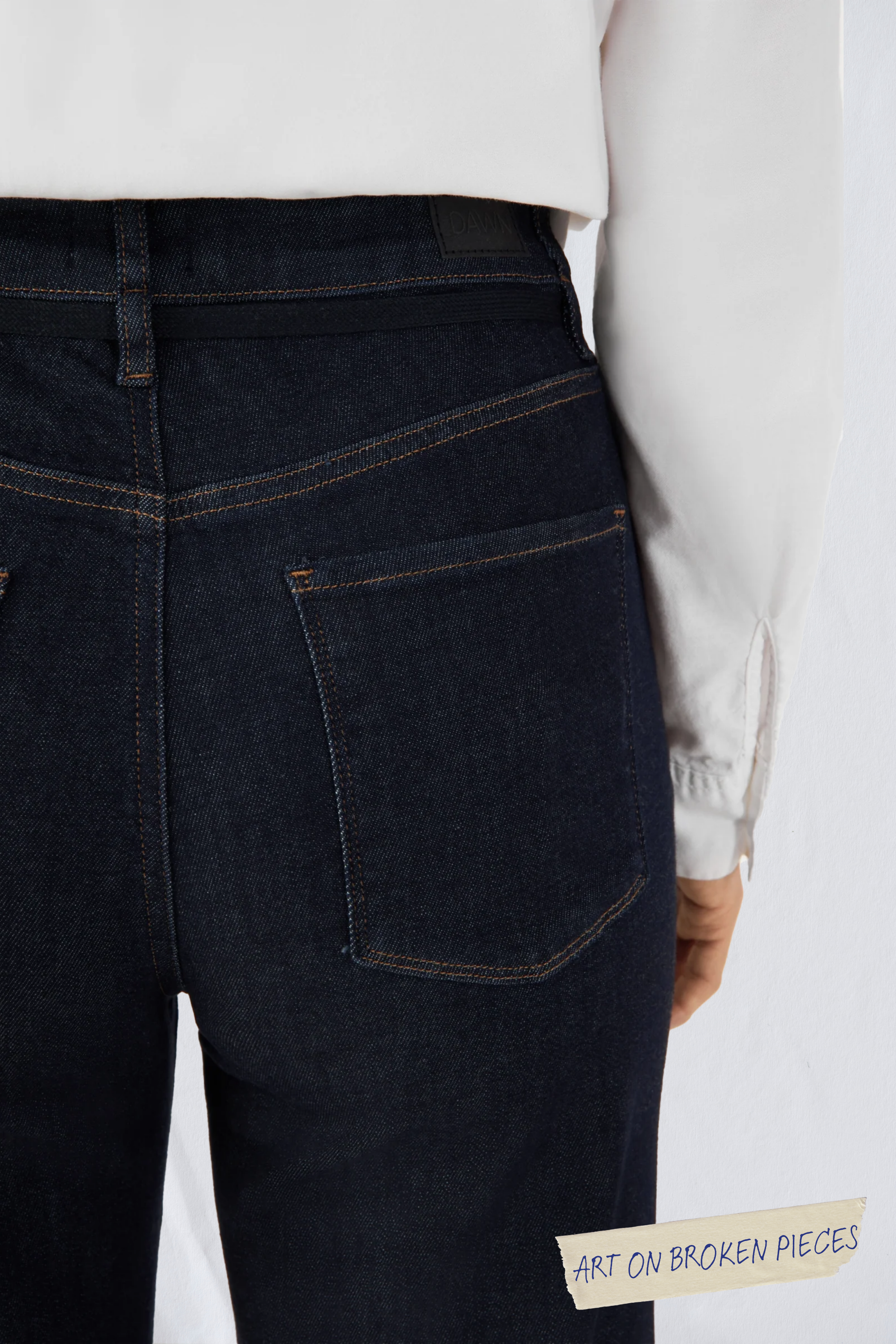 Rückansicht DEW Cropped Flared Jeans in Raw Blue: hoher Bund, ausgestelltes Bein, Gesäßtaschen, orange Nähte; ART ON BROKEN PIECES unten rechts.