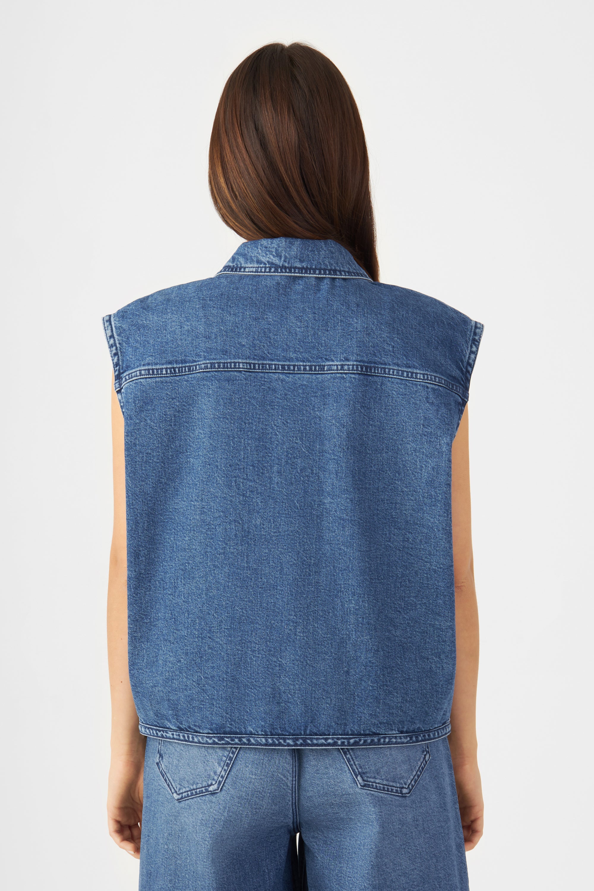 Rückansicht einer mittelblauen Denim-Weste im Biker-Stil, ärmellos, overschnittene Schultern, kurzer Schnitt.