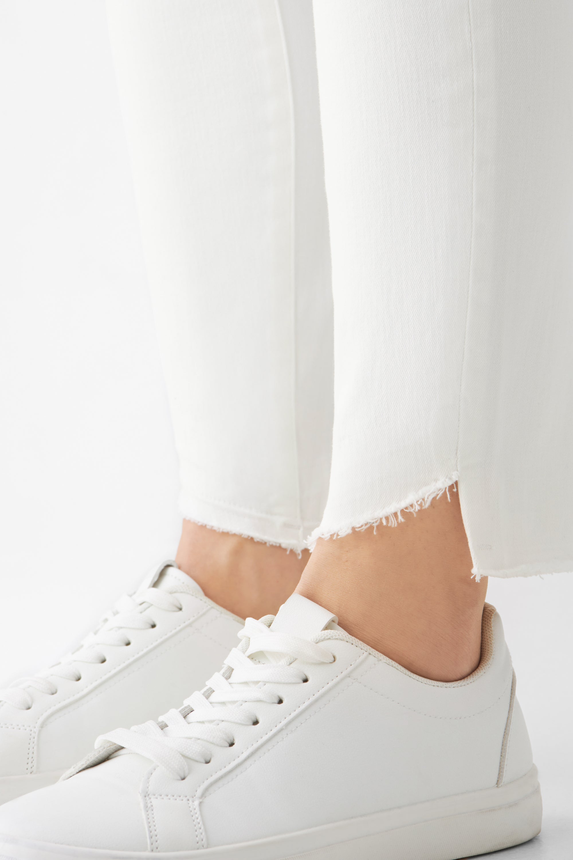 Weiße Slim-Fit Jeans aus Organic Comfortstretch mit offenem, fransigem Saum am Knöchel; kombiniert mit weißen Sneakern.
