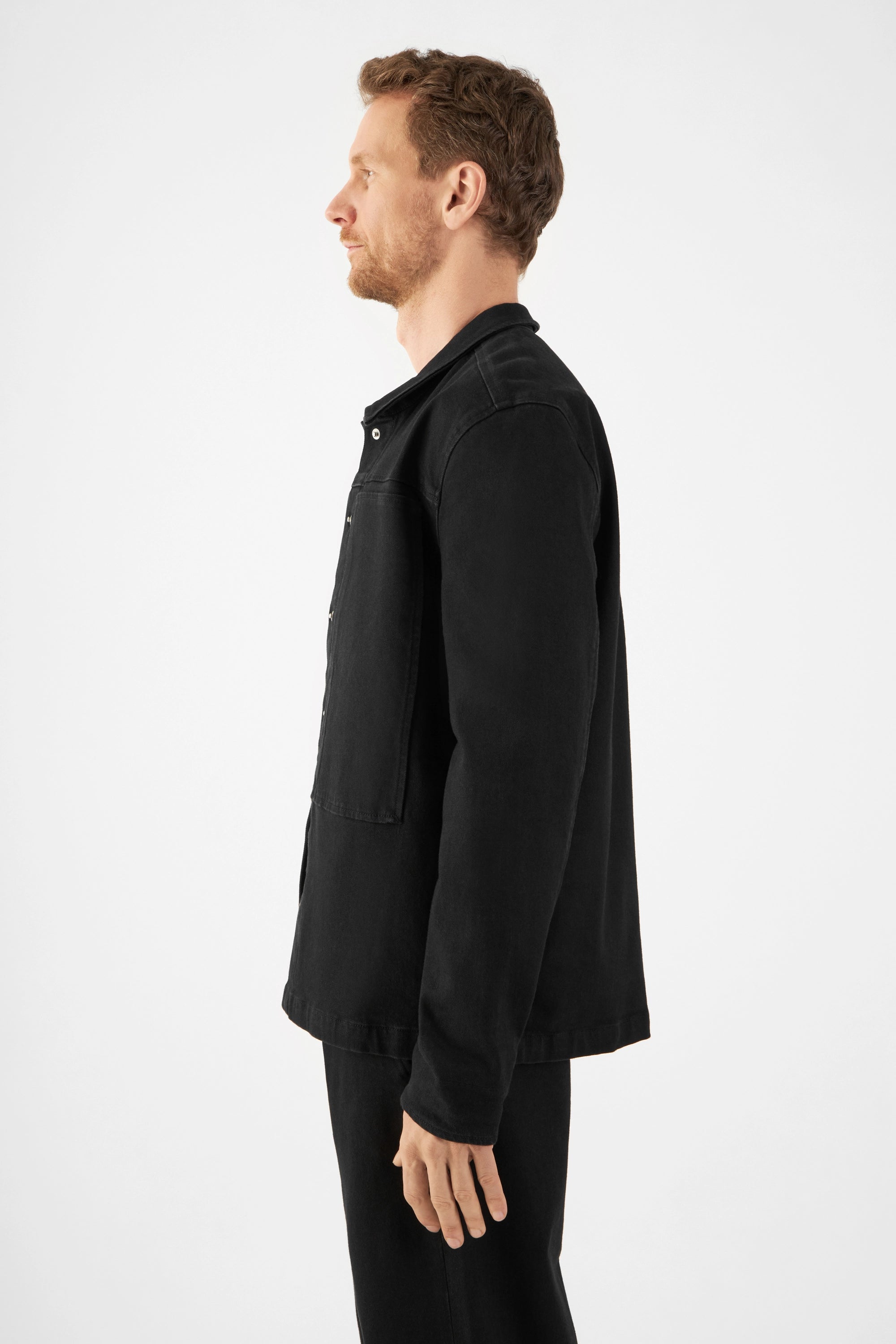 Seitenansicht eines Mannes in schwarzer QUIET Jacke aus Soft Denim TENCEL™ – kastiger, lockerer Schnitt, frontale Druckknöpfe.