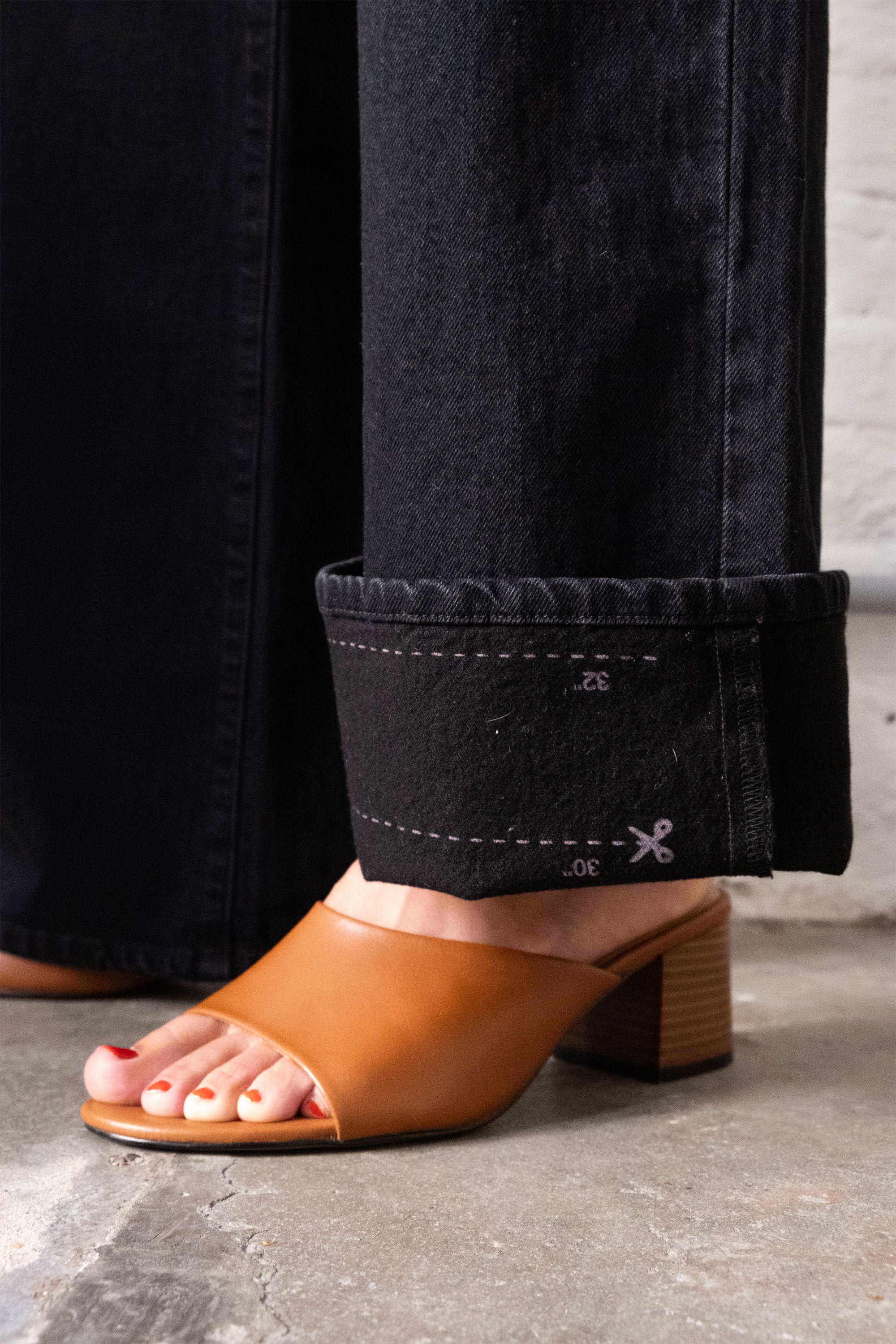 Schwarze Wide-Leg Jeans mit umgekrempeltem Saum; Saum-Markierungen am Saum, getragen zu cognacfarbenen Sandalen mit Blockabsatz.