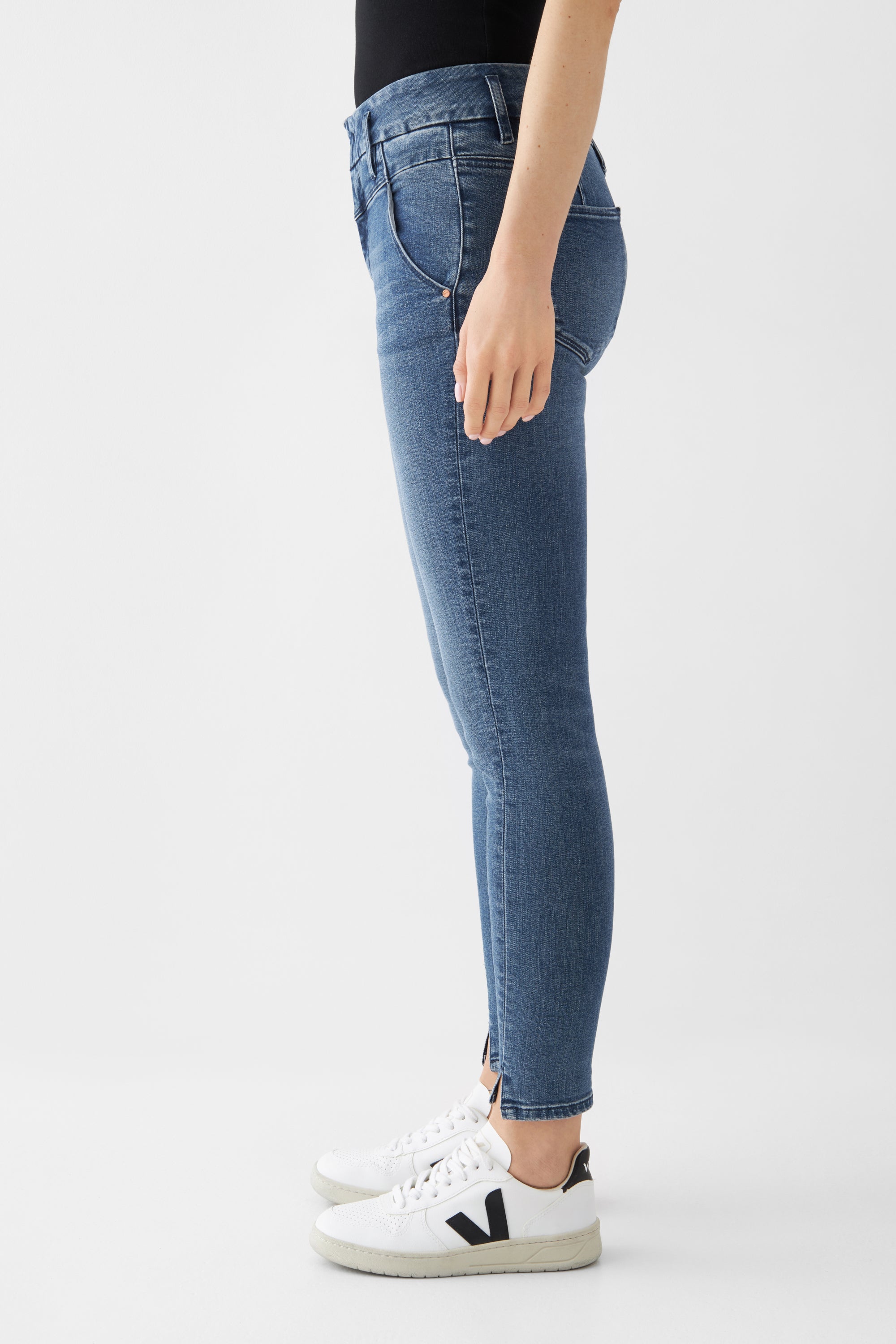 Sun Up Skinny Jeans in Medium Blue, seitliche Ansicht: schräge Außennähte und Saumschlitz; figurbetont durch Powerstretch, mit weißen Sneakern.
