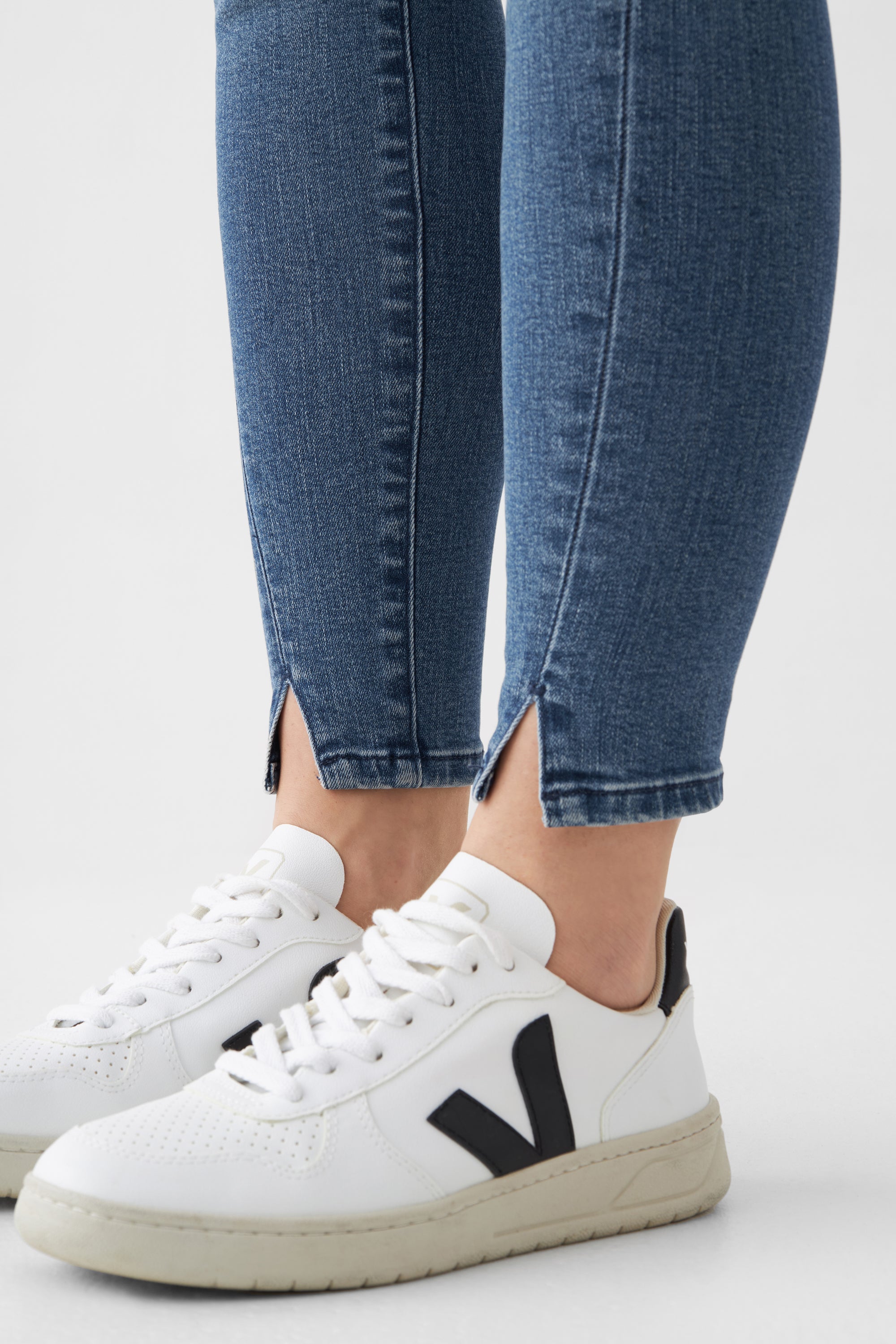 SUN UP Skinny Jeans in Medium Blue mit Powerstretch; schräge Außennähte und Saumschlitz; verkürztes Bein, getragen mit weißen Sneakern.