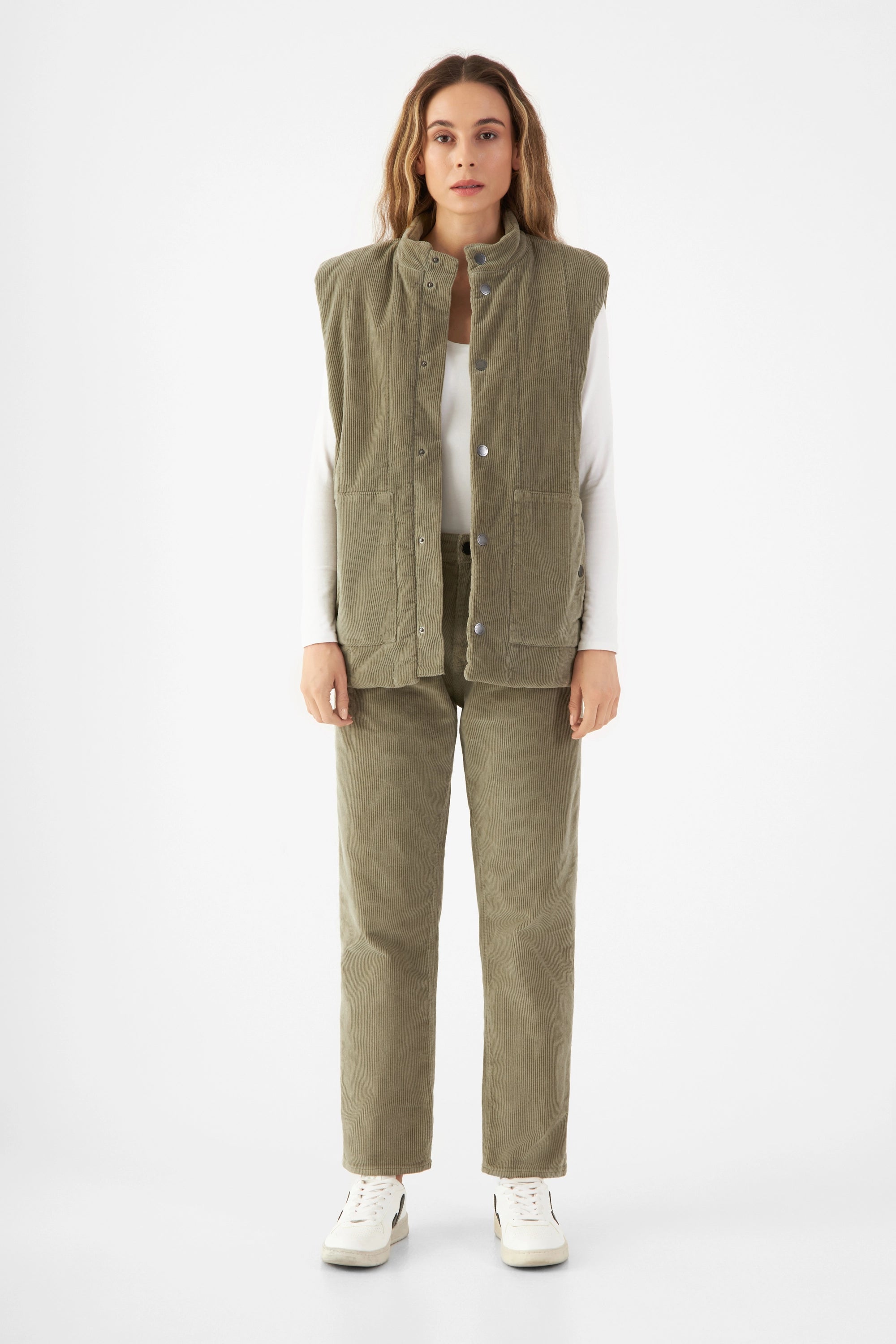KASHEW gepaddete Corduroy-Weste in Sage Green, vertikale Steppung, kastiger Schnitt, endet knapp über der Hüfte; getragen über weißem Langarmshirt.
