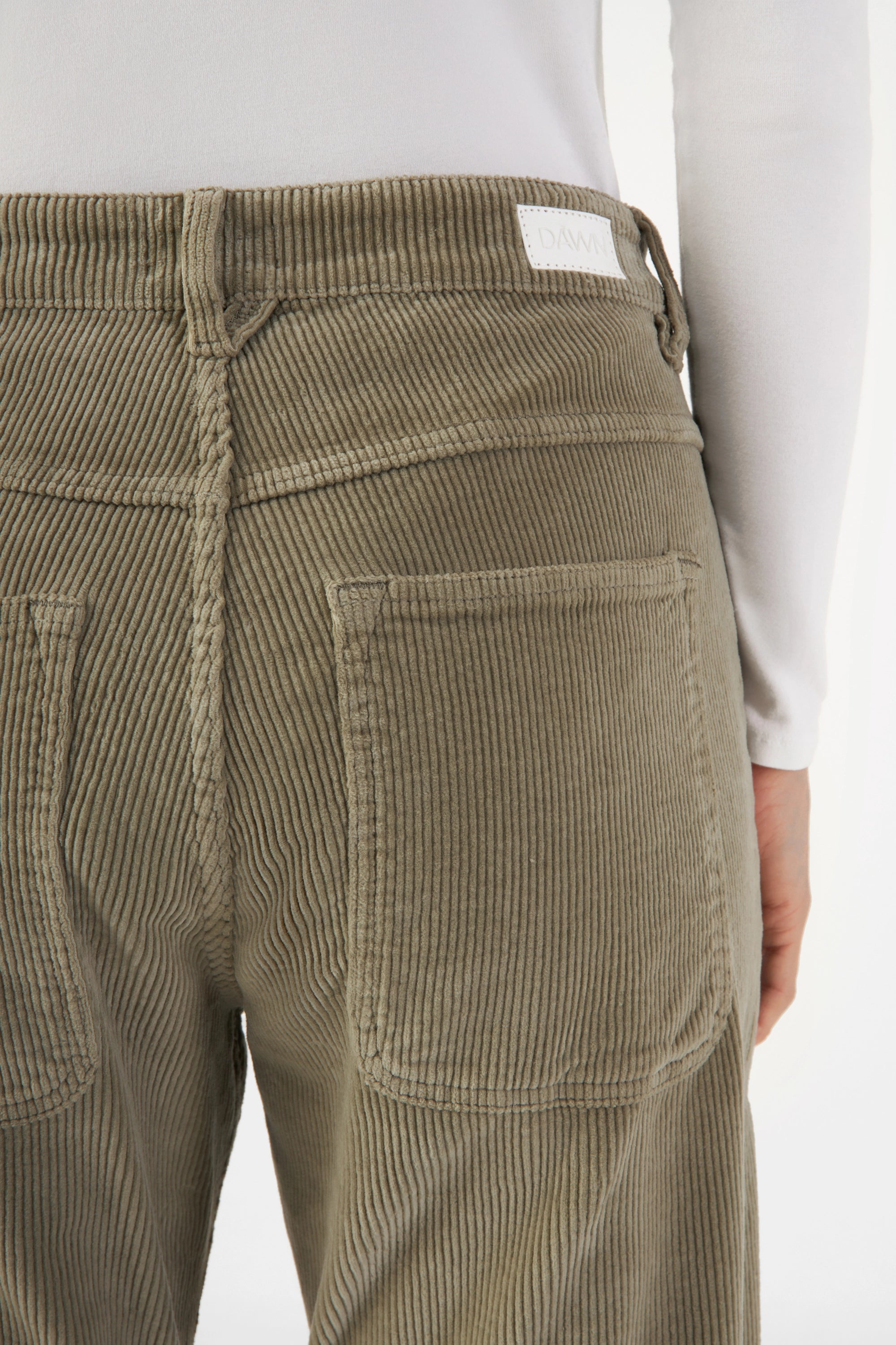 Sagegrüne MANE Corduroy-Hose, Rückenansicht mit Rückentasche, mittlerer Bund, feine Corduroy-Rippen, DAWN-Label am Bund.