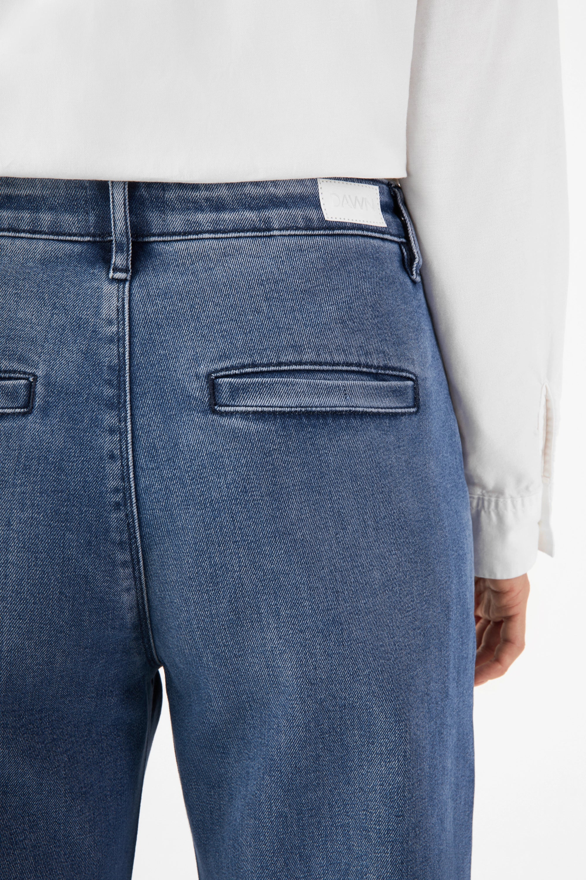 Rückansicht einer Denim-Chino in Medium Blue (GLOW) mit zwei hinteren Paspeltaschen; Bund mit Gürtelschlaufen und Markenlabel.