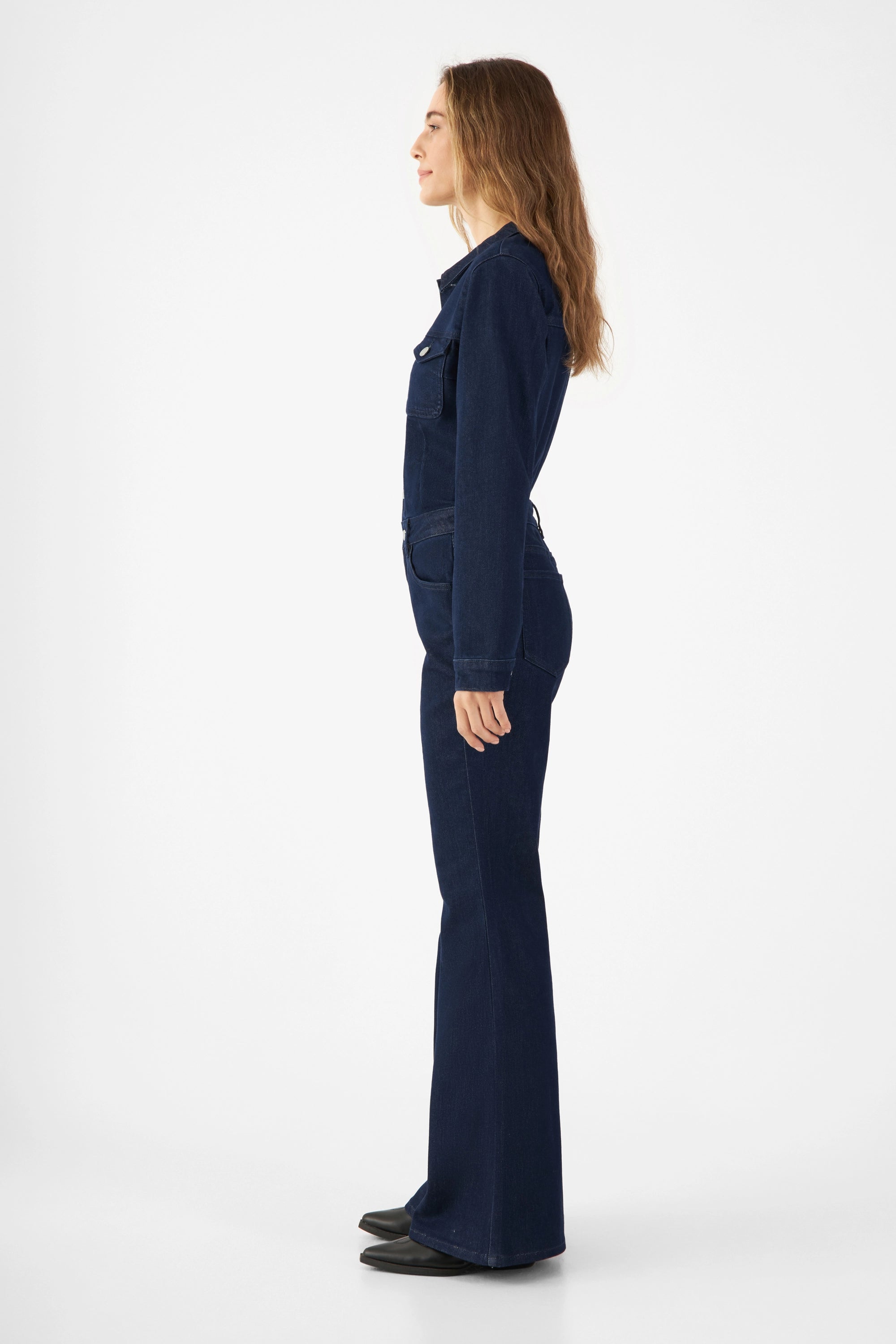 Seitliche Profilaufnahme eines Models in dunkelblauem GRACE Denim-Overall (Rinse Blue) mit langem Arm, tailliertem Oberteil und ausgestelltem Beinverlauf.