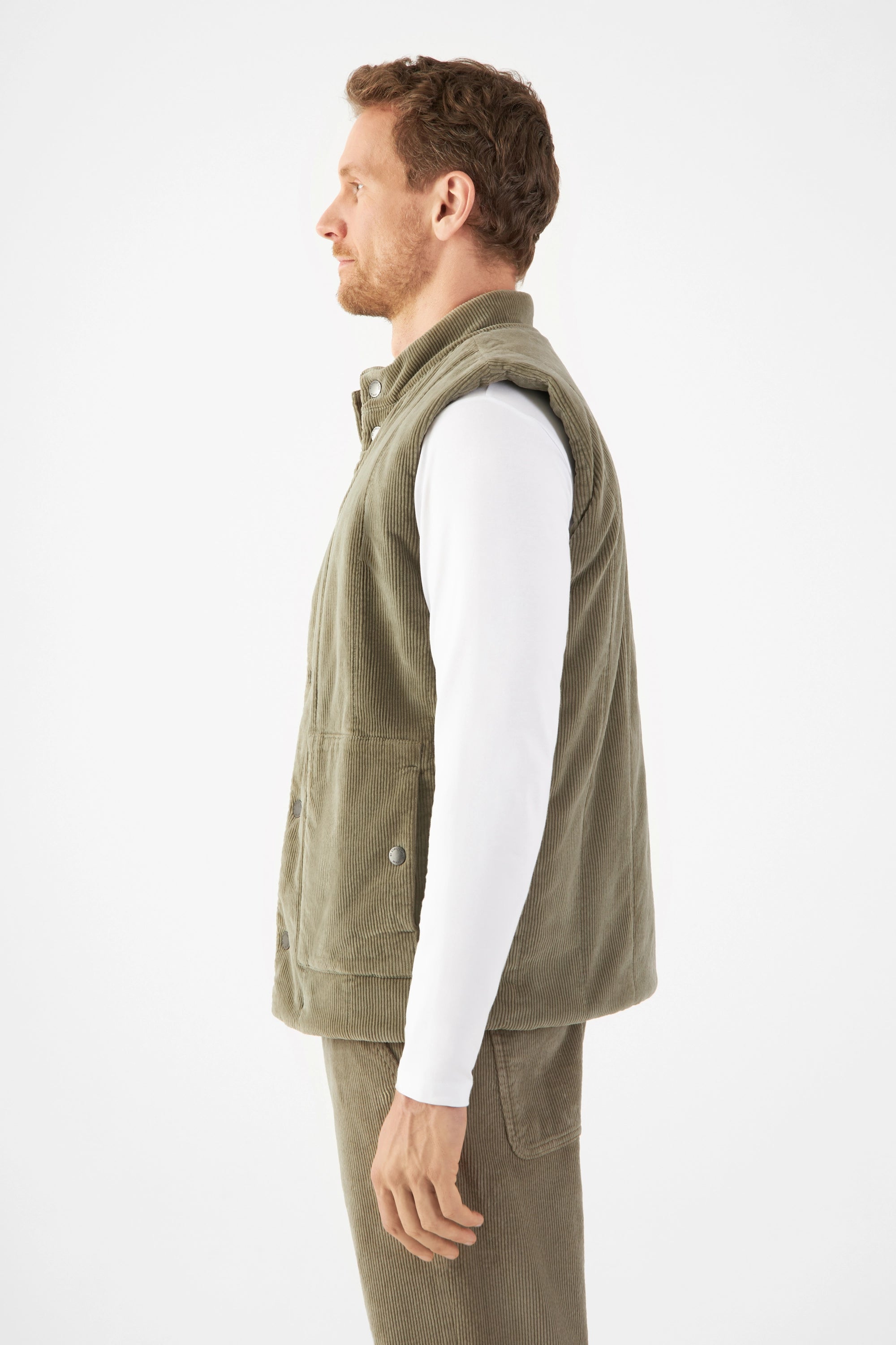 Seitenansicht eines Mannes in weißem Langarmshirt mit Sagegrün wattierter Corduroy-Weste (KASHEW); lockerer, kastiger Schnitt.