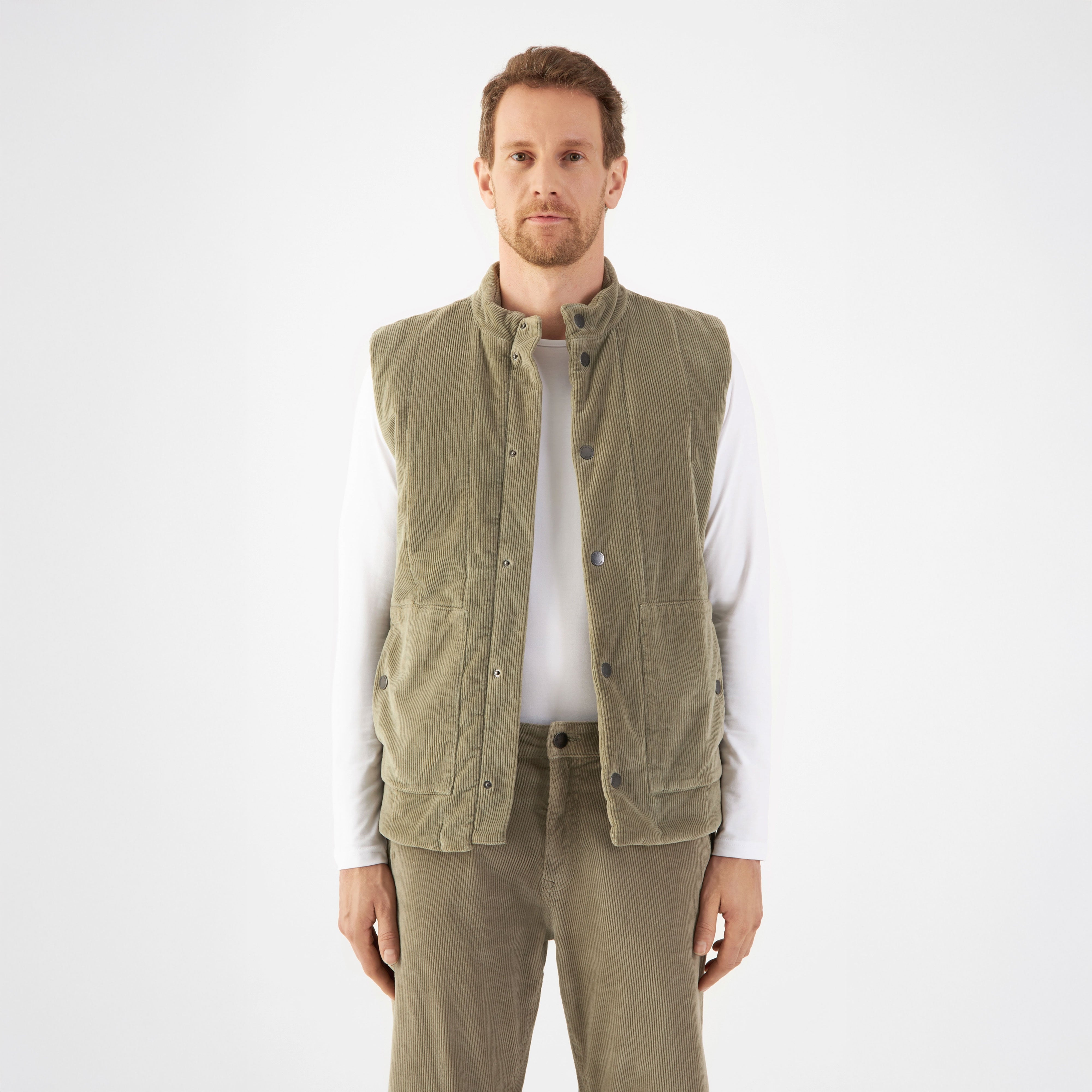Sagegrüne wattierte Corduroy-Weste KASHEW im Utility-Stil, vertikal gesteppt, lockerer kastiger Schnitt endet knapp über der Hüfte; getragen über weißem Langarmshirt, ideal zum Layern.