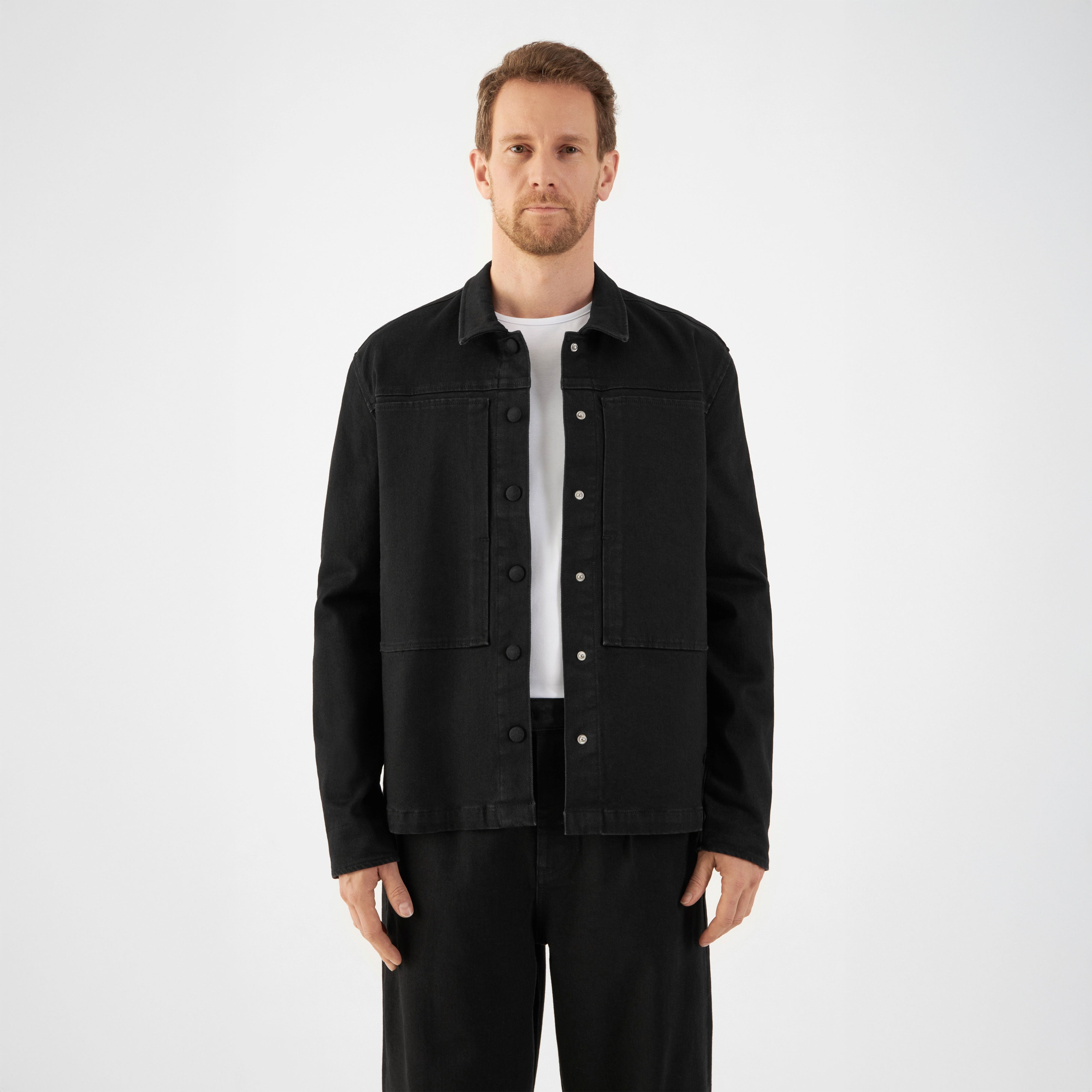 Schwarze QUIET-Jacke aus TENCEL-Denim, kastiger, entspannter Schnitt mit zwei Brusttaschen; Frontverschluss mit Druckknöpfen; vollständig gefüttert, getragen von einem Mann mit weißem T‑Shirt.